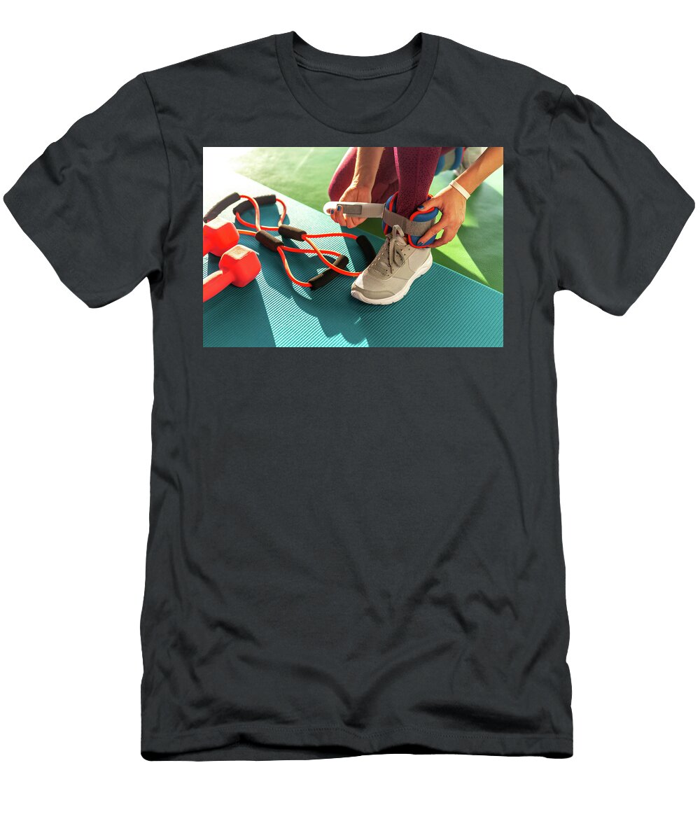 1 Woman Putting on Ankle Wights Before She Starts a Workout in a Gym Health and Fitness Concept with Dumbbells on Mat Maria Kray T-Shirt